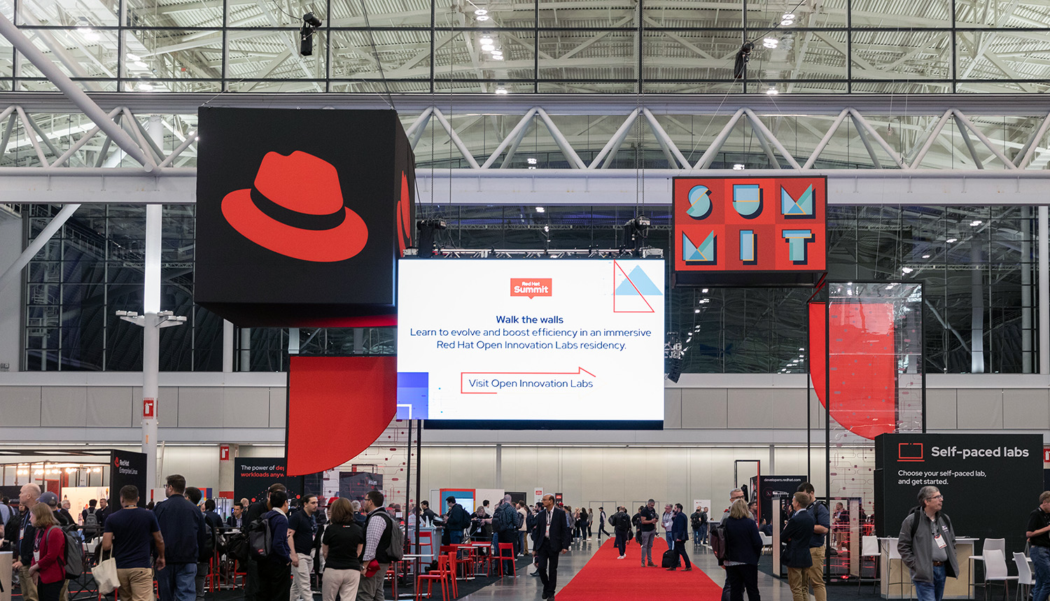 A photo in the expo hall at Red Hat Summit showing a large sign with the hat hanging above the booths.