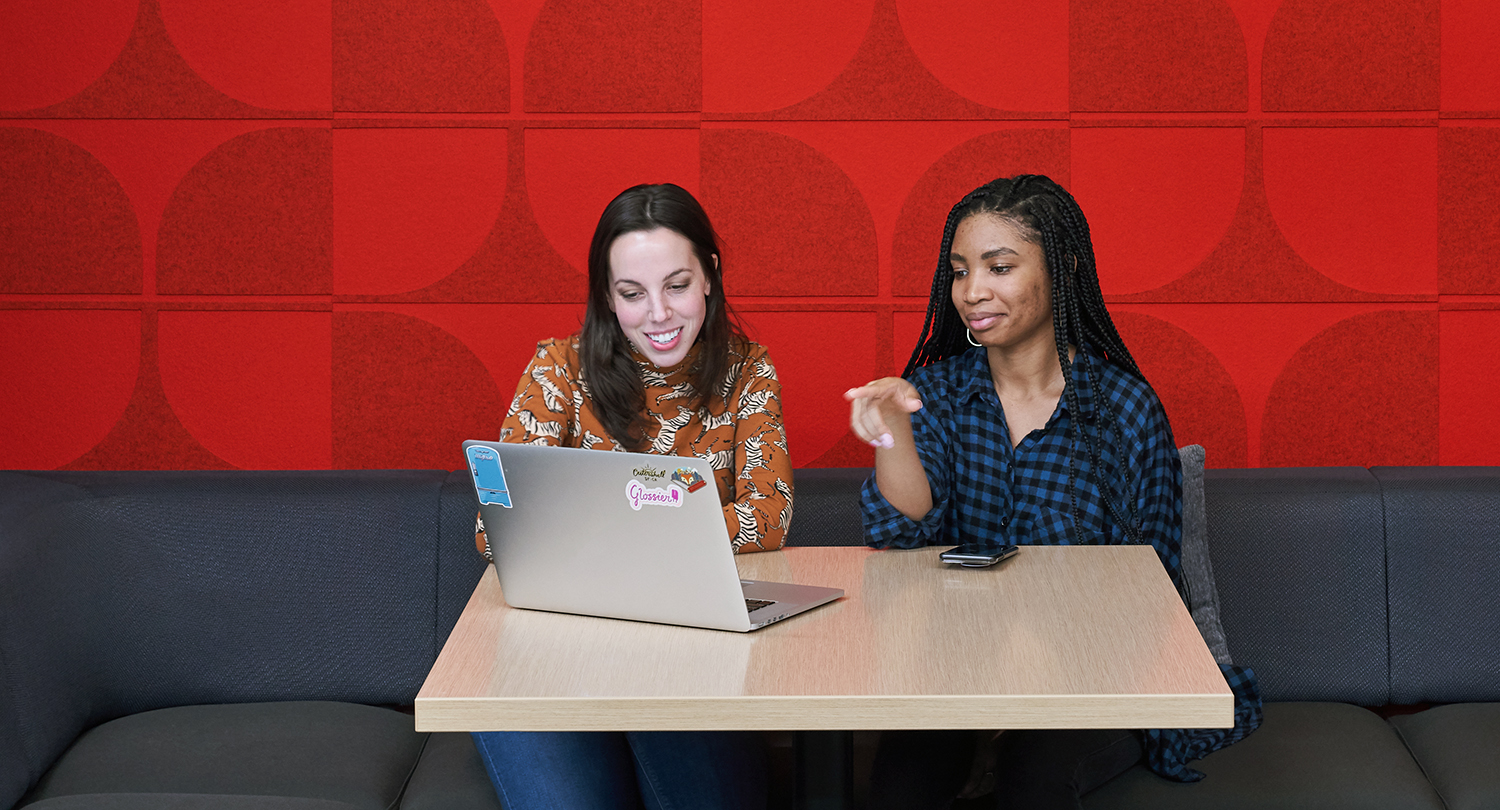 Red Hatters collaborate at a table at Red Hat Tower. Behind them is a wall made of red felt with a pattern reminiscent of the curves of a fedora.