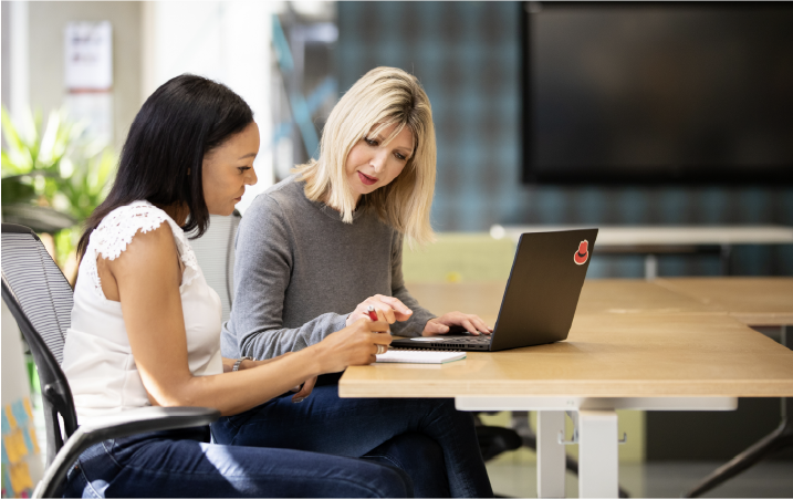 A custom stock photo taken showing two Red Hatters sitting together at a table, working on a laptop that has a Red Hat sticker.