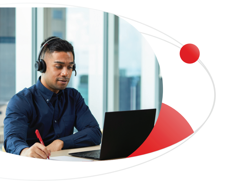 A man wearing a headset is on a video call while taking notes at his desk in a modern office setting.